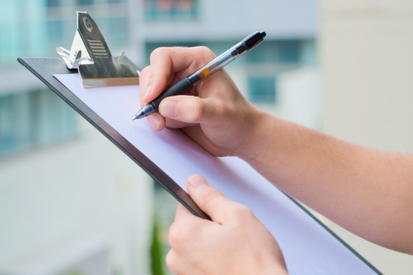 cropped view of an inspector's hand writing on paper on clipboard depicting oil tank compliance checks